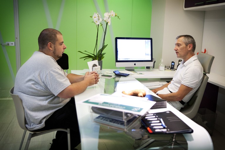 A patient with a higher body weight meets with their physician in a weight‑management clinic, speaking across a desk in a private office setting.