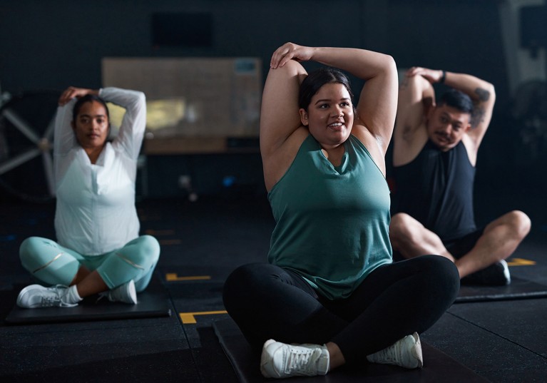 Three higher-weight people are stretching while sitting down on mats during a workout at a gym.
