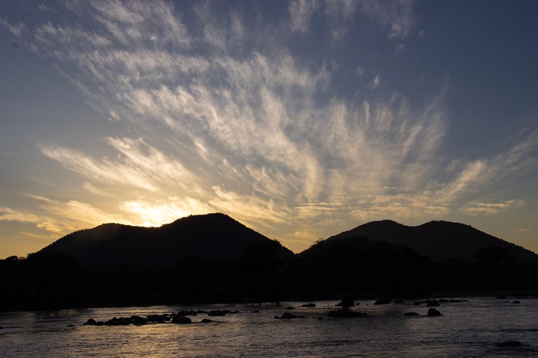 High-altitude cirrus clouds are illuminated by the first light of day above two mountains, with a lake in the foreground.