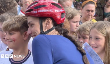 Scott stands in cycling gear and helmet with children and adults who have come to see her on her challenge. She is posing for photos with children who are smiling. She has long brown hair in a plait, emerging from under a red cycle helmet, and is wearing a blue cycle top.