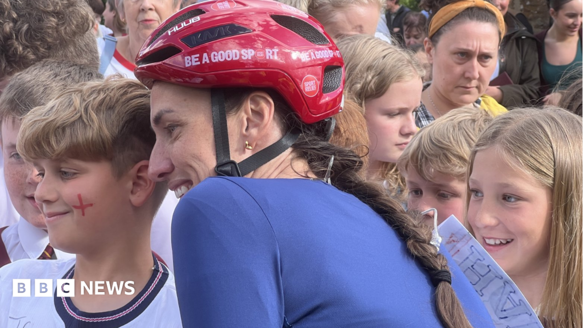 Scott stands in cycling gear and helmet with children and adults who have come to see her on her challenge. She is posing for photos with children who are smiling. She has long brown hair in a plait, emerging from under a red cycle helmet, and is wearing a blue cycle top.