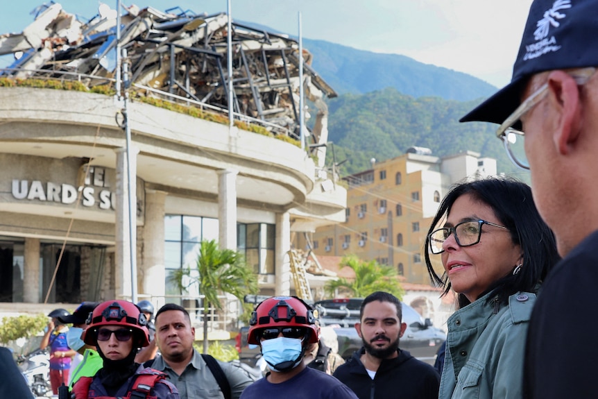 a woman with short balck hair, black glasses is surrounded by officials at a damaged building.