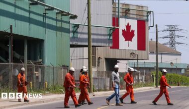 A Canadian national flag is seen in the background as seven workers dressed in orange work suits and white hard hats cross the street in front of ArcelorMittal Dofasco's steel manufacturing buildings in Hamilton, Ontario.