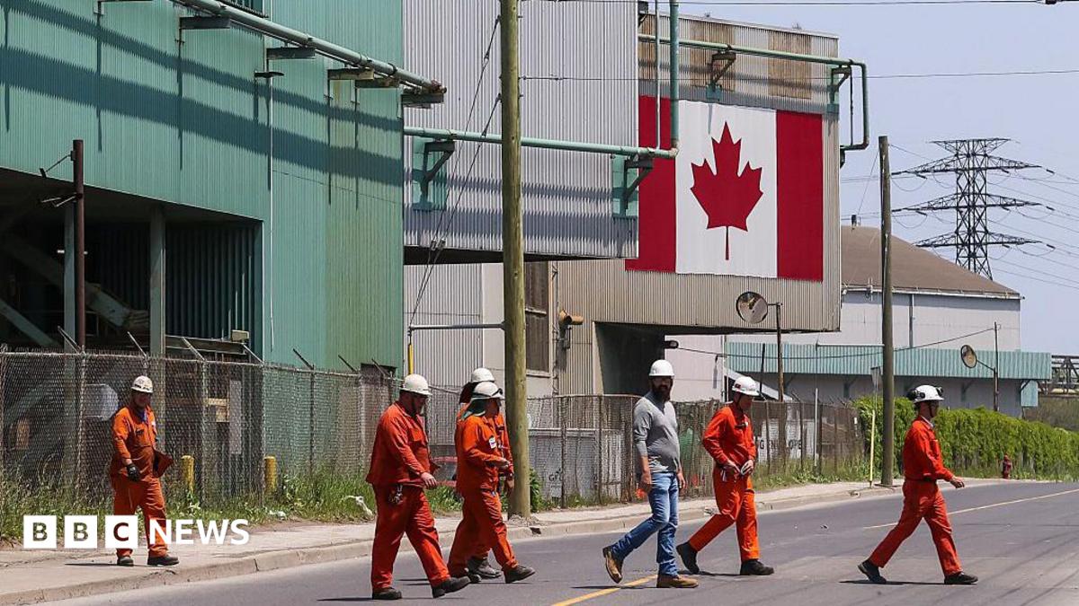 A Canadian national flag is seen in the background as seven workers dressed in orange work suits and white hard hats cross the street in front of ArcelorMittal Dofasco's steel manufacturing buildings in Hamilton, Ontario.