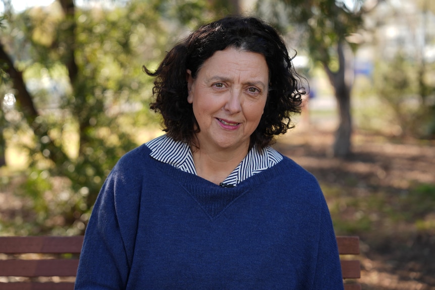 A woman with dark hair with a blue and white striped collar protruding from a blue jumper stares at the camera in a park.