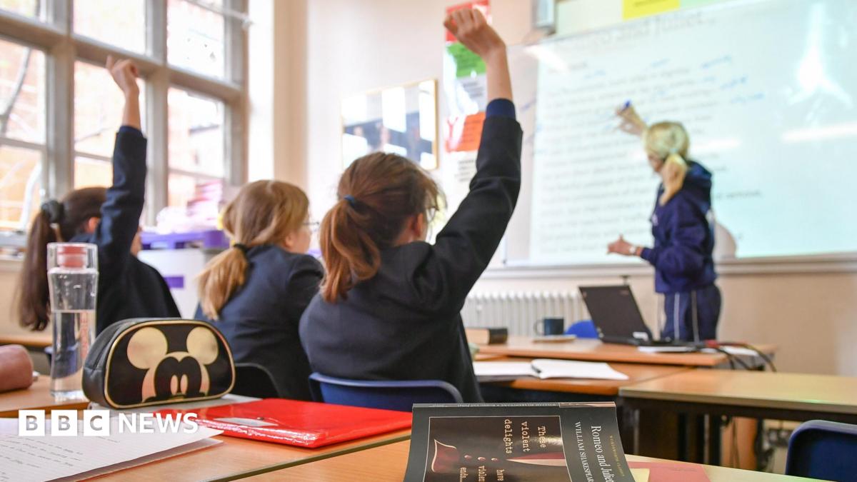 Three female students sitting in a classroom with their back to the camera while a female teacher writes on a board in the distance. The pupils are wearing school uniform and two have their right hands raised.