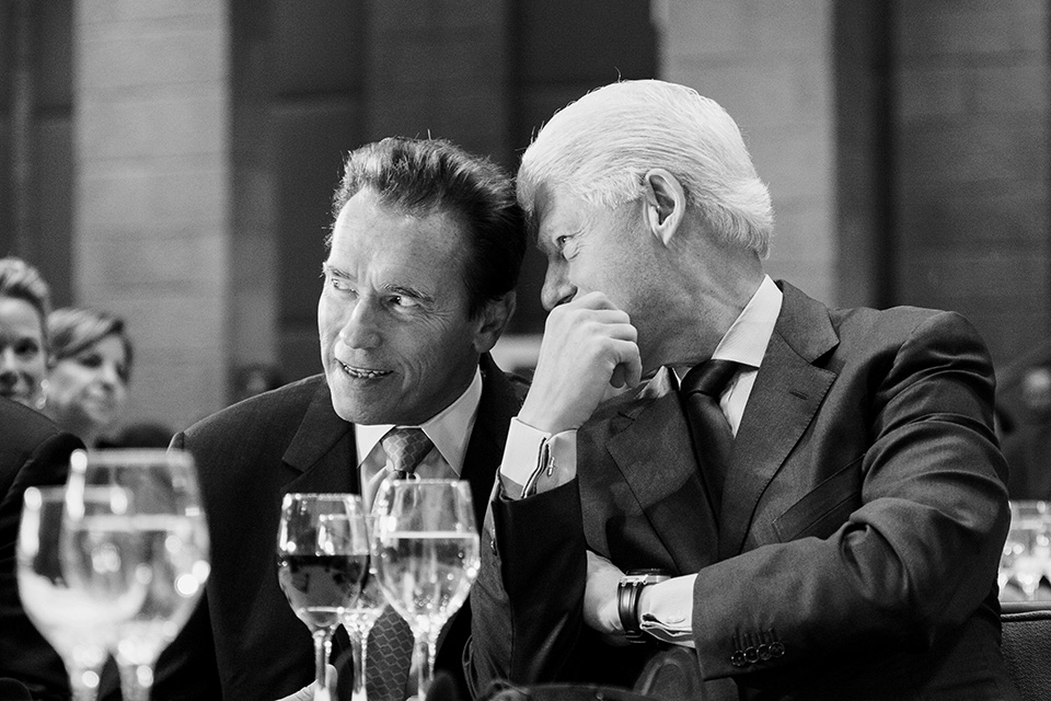 A candid black-and-white shot of Schwarzenegger leaning in to share a private word with a silver-haired Bill Clinton in at a formal dinner, glasses and wine visible on the table in front of them.