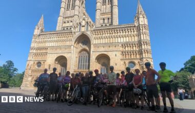 A group of cyclists in cycling gear with bikes stood in front of Lincoln Minster. The sky is blue.