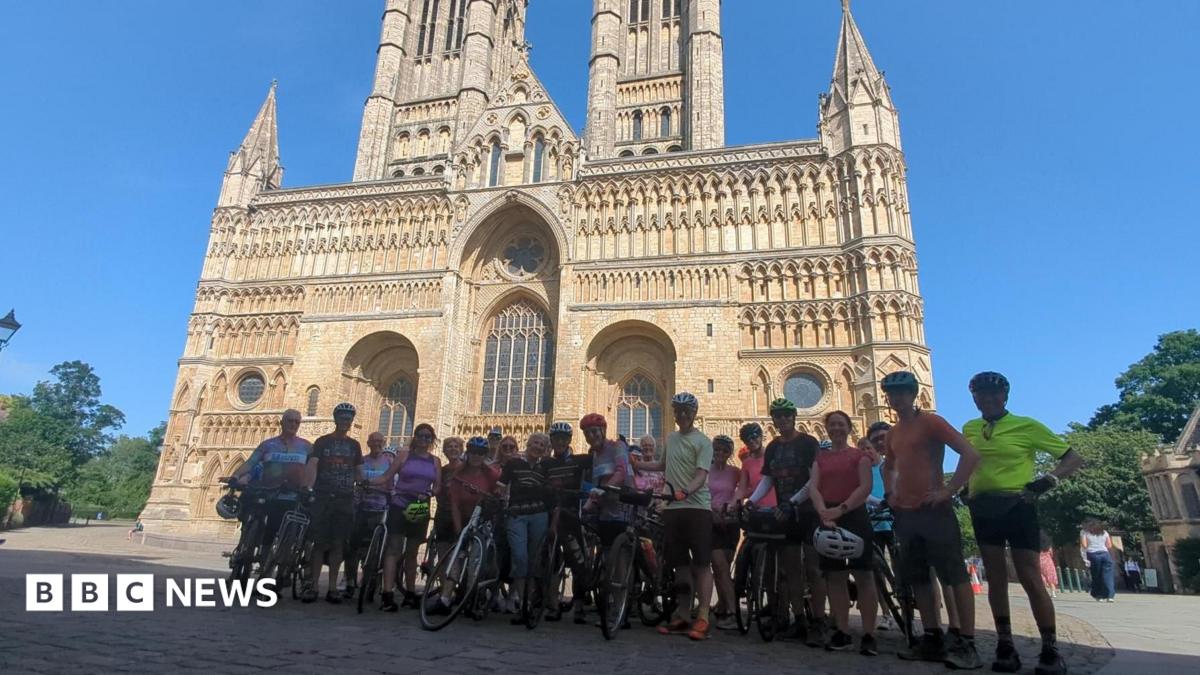 A group of cyclists in cycling gear with bikes stood in front of Lincoln Minster. The sky is blue.