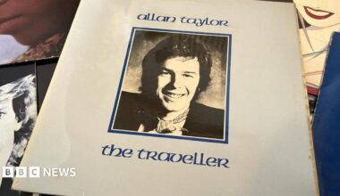 Allan Taylor seated and looking at the camera and smiling with a guitar o his lap and both arms resting on it.