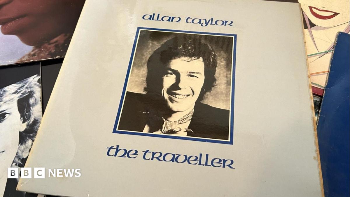Allan Taylor seated and looking at the camera and smiling with a guitar o his lap and both arms resting on it.