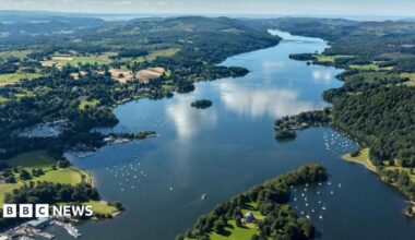 An aerial view of Windermere, England's largest lake which stretches into the distance narrowing as it goes. Boats can be seen dotted along its foreshore