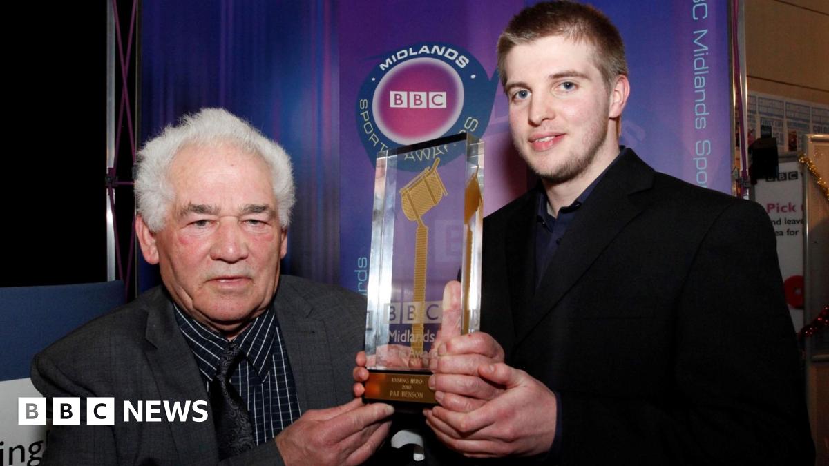 Two people standing indoors at an event booth display branded ‘BBC Midlands’ in the background. Both individuals are holding a tall, transparent trophy featuring a golden microphone design mounted on a base with engraved text. The backdrop includes a purple gradient panel with a circular BBC Sports logo and vertical ‘BBC Midlands’ text along the side. The setting appears to be an awards or media event, with exhibition stands and signage visible in the background.