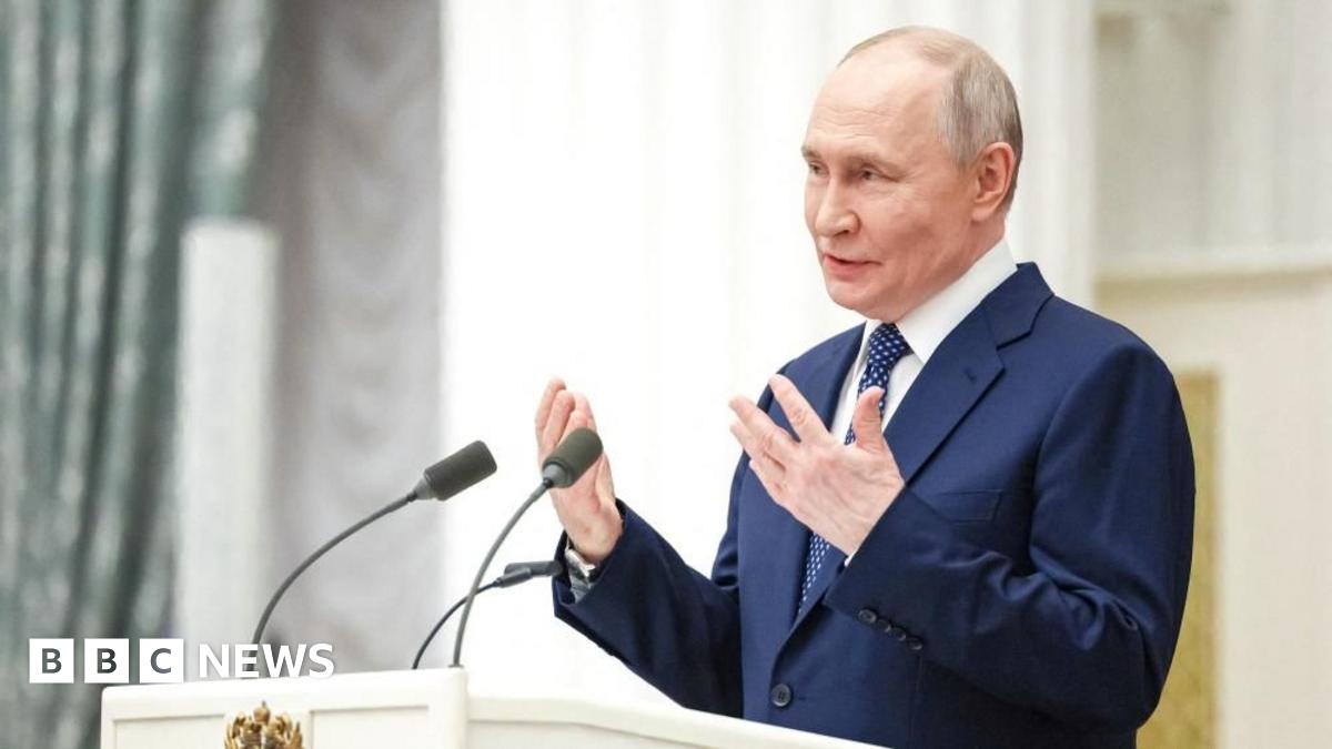 President Putin wearing a white shirt, dotted blue tie and blue suit, gestures with his hands as he speaks at a white podium with two microphones. A light green patterned curtain stands to the left of the picture.