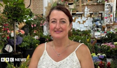 There is a woman who is wearing a spaghetti lace white t-shirt and a pendant. She has dark hair tied up. Behind her is lots of colourful flowers and plants, plus a big noteboard over her left shoulder.