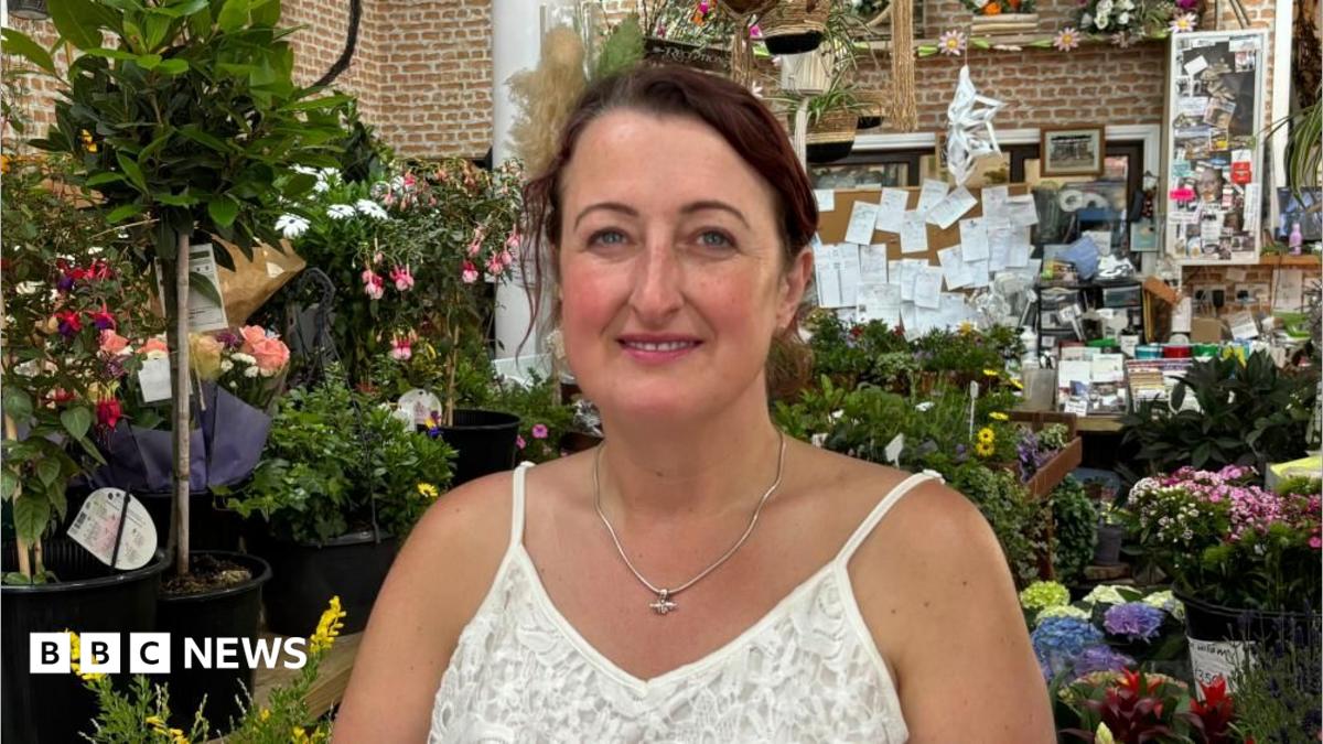 There is a woman who is wearing a spaghetti lace white t-shirt and a pendant. She has dark hair tied up. Behind her is lots of colourful flowers and plants, plus a big noteboard over her left shoulder.