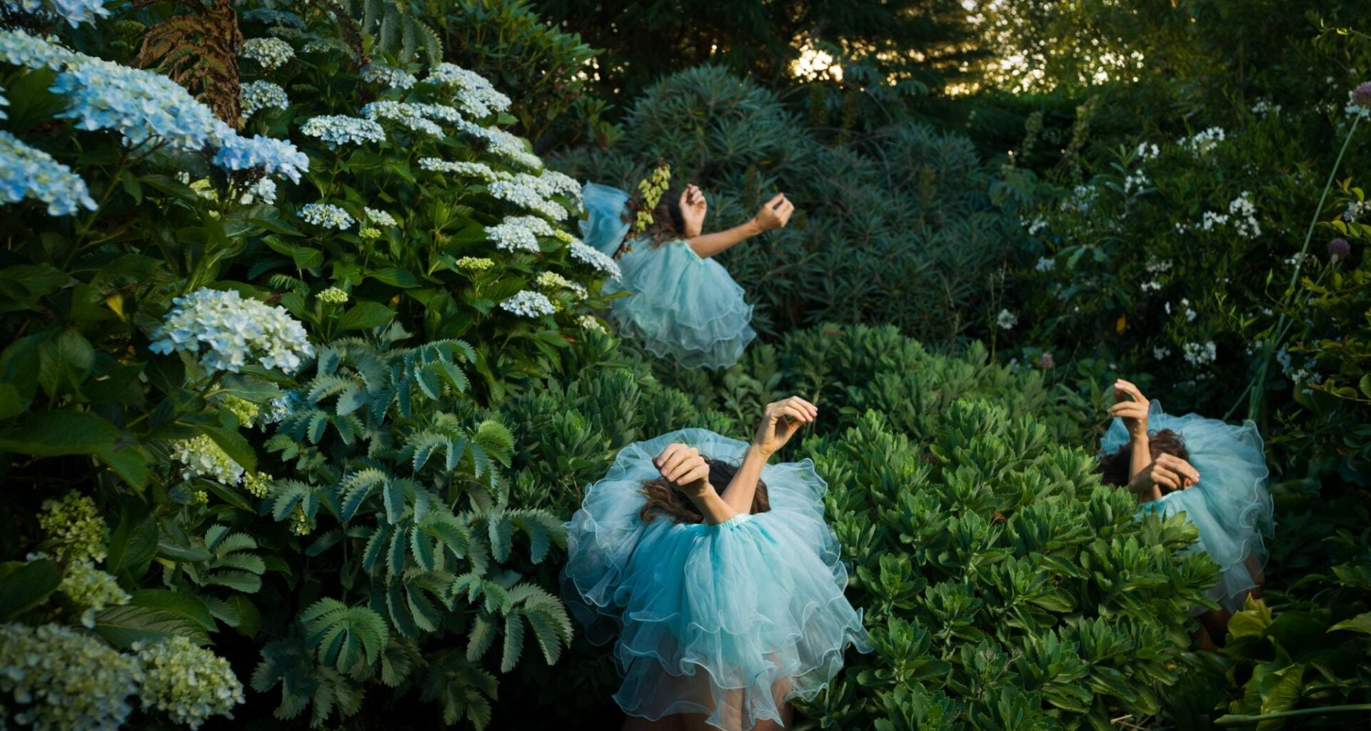 a photograph by Tamara Dean of three individuals sitting in a dense brush, wearing blue tutus around their bodies, only their arms sticking out