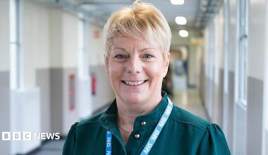 Michelle Arrowsmith, a woman with blonde short hair standing in a hospital corridor. She is looking directly at the camera and smiling and is wearing a dark green blouse and blue NHS lanyard round her neck.