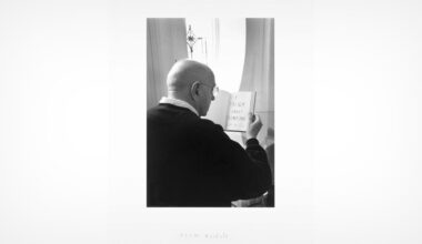 A bald person wearing glasses reads a small book with the words "THINK ABOUT SOMETHING" written on the open page, standing near a window with light streaming in.