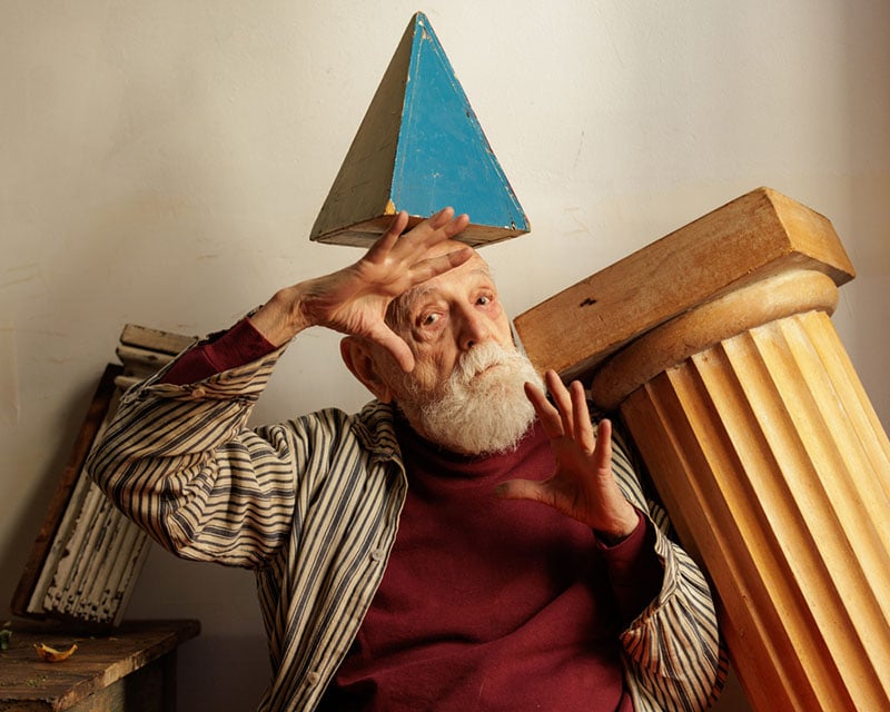 An elderly man with a white beard sits indoors, balancing a blue triangular prism on his head. He gestures with his hands and leans against a large, toppled column. He's dressed in a striped shirt over a maroon top.