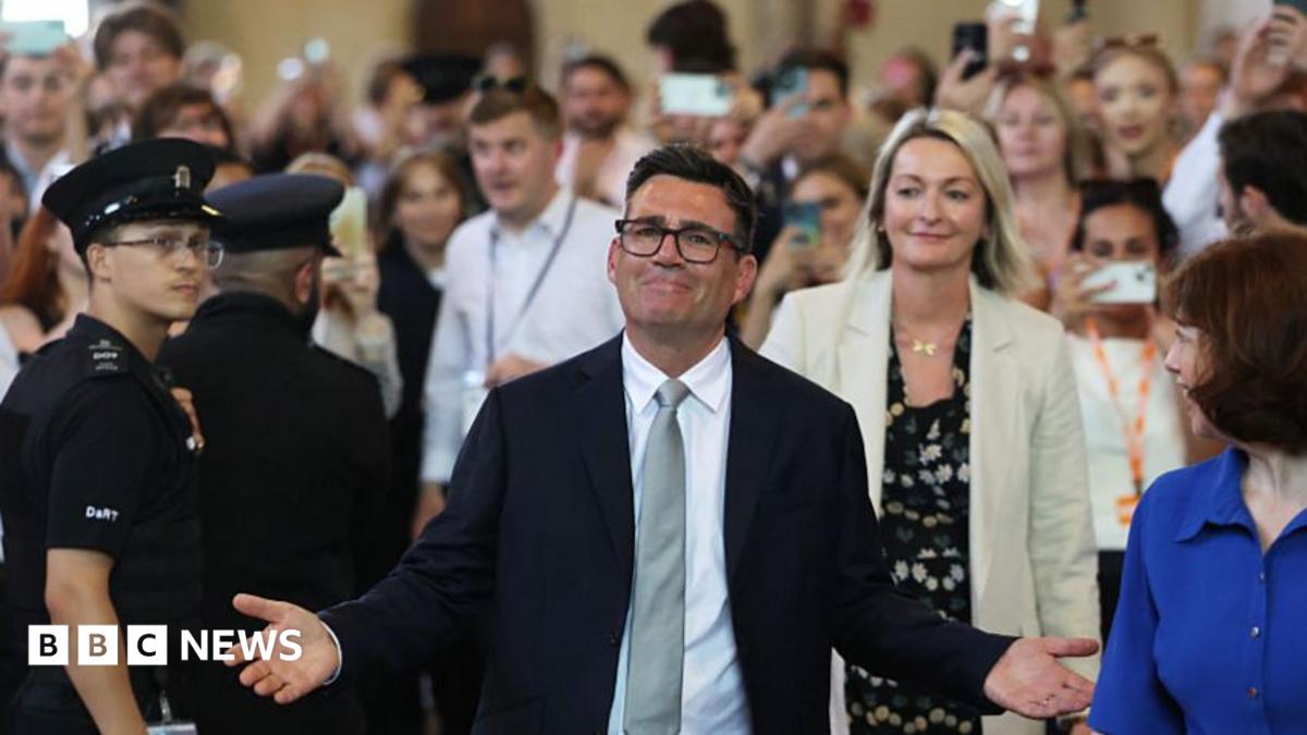 Andy Burnham, wearing a dark suit, silver tie and white, with arms open and a crowd behind him