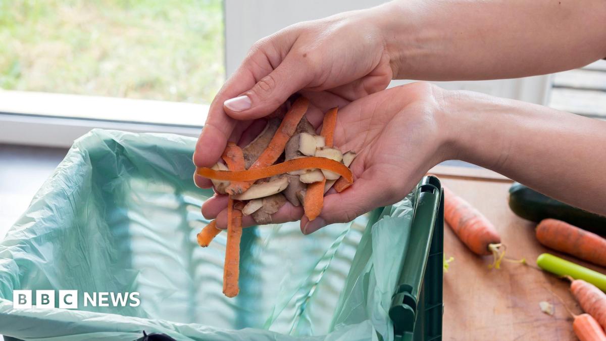 A pair of hands drops carrot and potato peelings into a green food waste caddy lined with a compostable bag. Whole carrots and a courgette sit on a wooden chopping board beside the caddy.