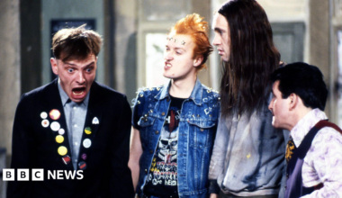 An image from sitcom The Young Ones, showing Mayall, Ade Edmondson, Planer and Christopher Ryan. Mayall, in character wearing a dark jacket including badges, is looking shocked and the others are looking at him.