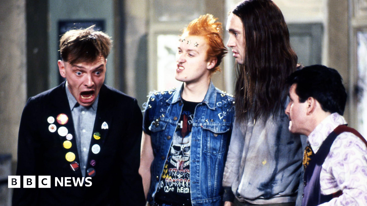 An image from sitcom The Young Ones, showing Mayall, Ade Edmondson, Planer and Christopher Ryan. Mayall, in character wearing a dark jacket including badges, is looking shocked and the others are looking at him.