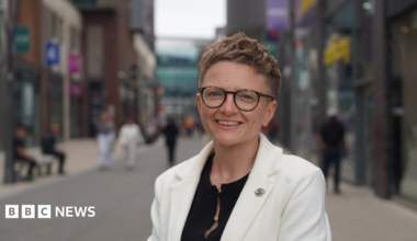 Bev Craig has short red hair, and is wearing glasses and a white jacket over a black blouse