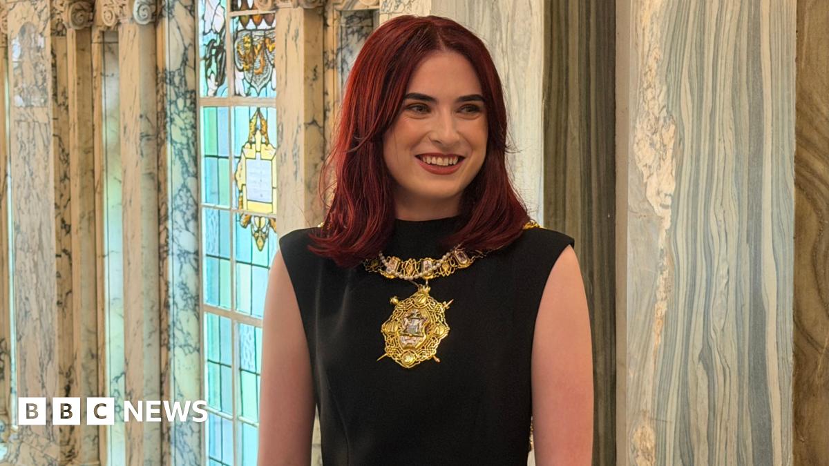 Róis-Máire Donnelly is smiling. She's wearing lipstick. She has red hair. She's wearing her gold chain of office and a black dress. A stained glass window is behind her and marble walls.