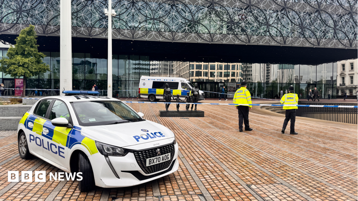 A police car parked on a paved area in front of a large building with a patterned metal front and a number of police officers in yellow jackets standing around