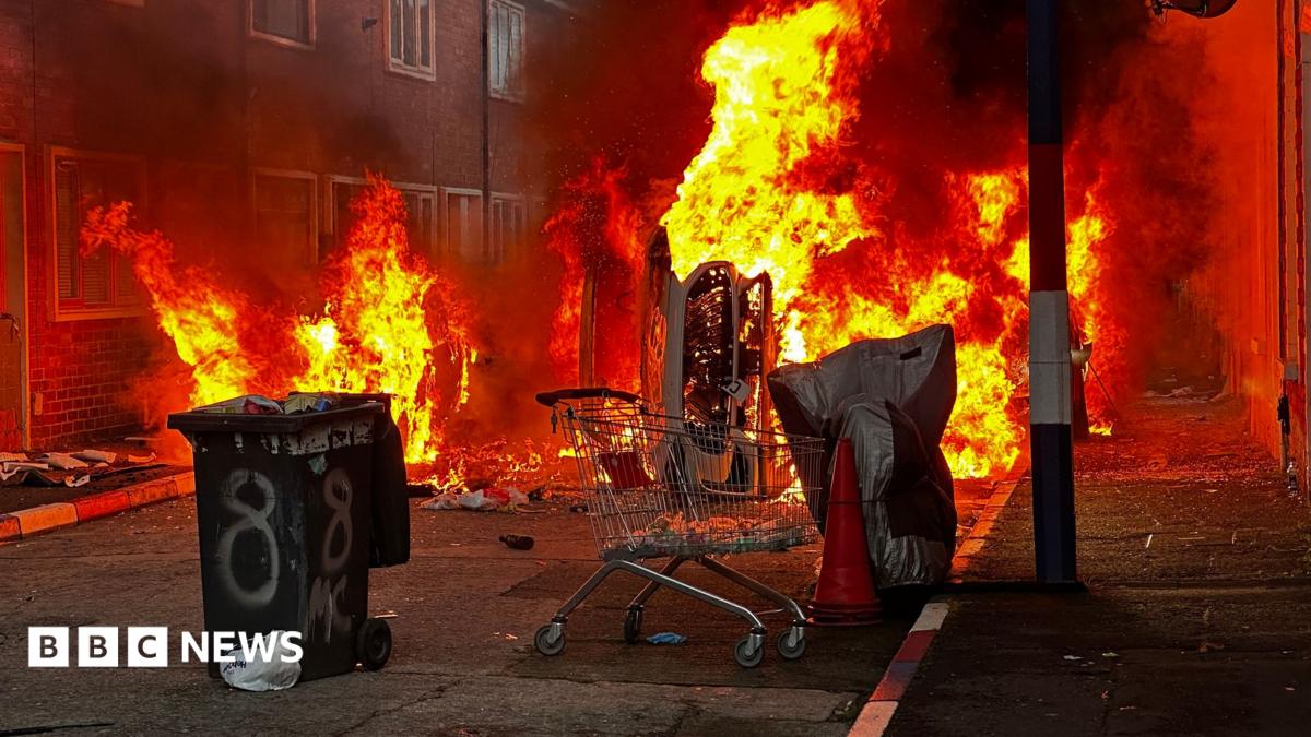 A wheelie bin and a trolley and a car are all on fire on a street. The kerb stones are painted red, white and blue.
