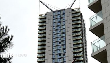 A tall, slender residential high-rise with distinctive V-shaped spires at the top, rising high above a line of green trees under an overcast sky.