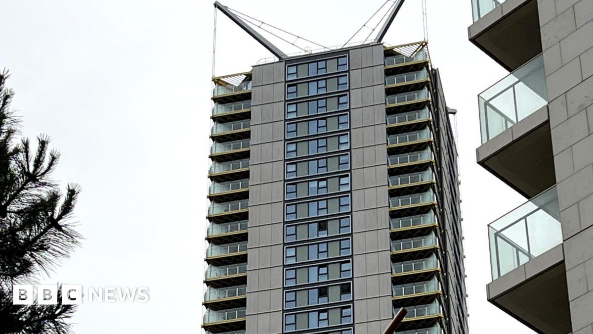 A tall, slender residential high-rise with distinctive V-shaped spires at the top, rising high above a line of green trees under an overcast sky.
