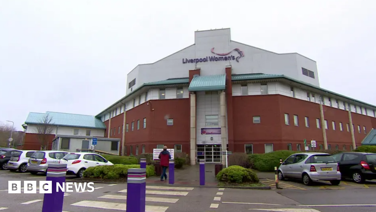 Exterior shot of Liverpool Women's Hospital, the main building is brown/orange, the roof is white. Liverpool Women's is written on the front in purple. Purple bollards lead up to the entrance.