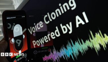 This picture is has a touchscreen mobile phone being held next to a screen which reads "voice cloning powered by AI".  The mobile phone screen display has a animated radio microphone with a red background. The lower third of the mobile phone screen has a record button and a timing display.