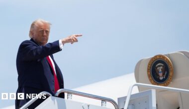 US President Donald Trump, at the top of a staircase up to Air Force One, pointing to the right. He is wearing a navy blue suit, white short and red neck tie.