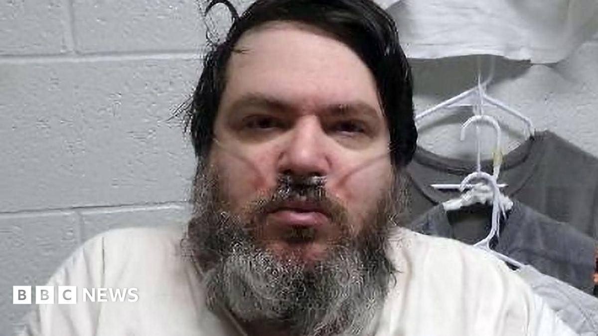 A man with black hair and a long black/grey beard has a breathing tube in his nose