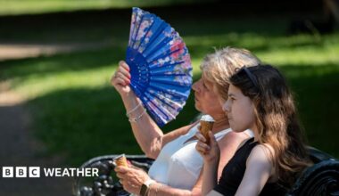 two women sitting on a park bench.  One eating an ice cream, the other fanning herself in the hot weather