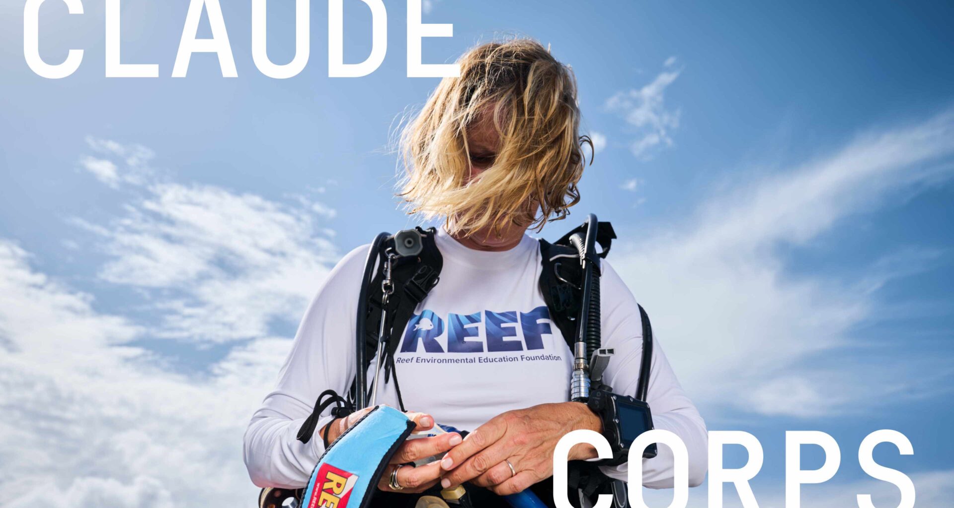 A diver in a long-sleeve white REEF shirt adjusts scuba gear against a bright blue, cloud-streaked sky.