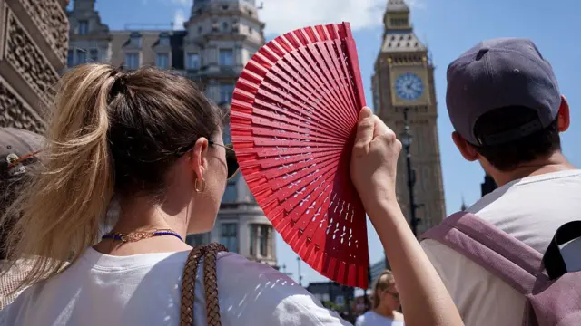 A visitor to the capital fans herself in front of Parliament with a red fan. London's Palace of Westminster is visible in the background.