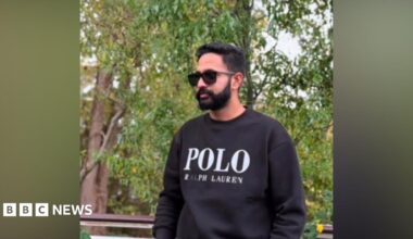 A young man standing outdoors near some trees. He is wearing a dark jumper and sunglasses and has a black beard and black hair.