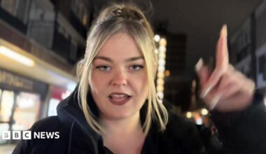 Morgan Da Savage has blonde long hair and is wearing a black top. She has her fingers up in a "gunfinger" position and smiles into the camera as she performs her rap in the Steeze Factory in front of a group of other people.