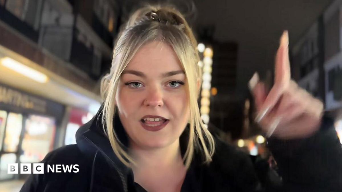 Morgan Da Savage has blonde long hair and is wearing a black top. She has her fingers up in a "gunfinger" position and smiles into the camera as she performs her rap in the Steeze Factory in front of a group of other people.