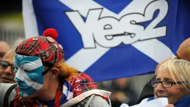 Man wearing a Saltire mask and tartan bunnet in front of a Yes2 flag