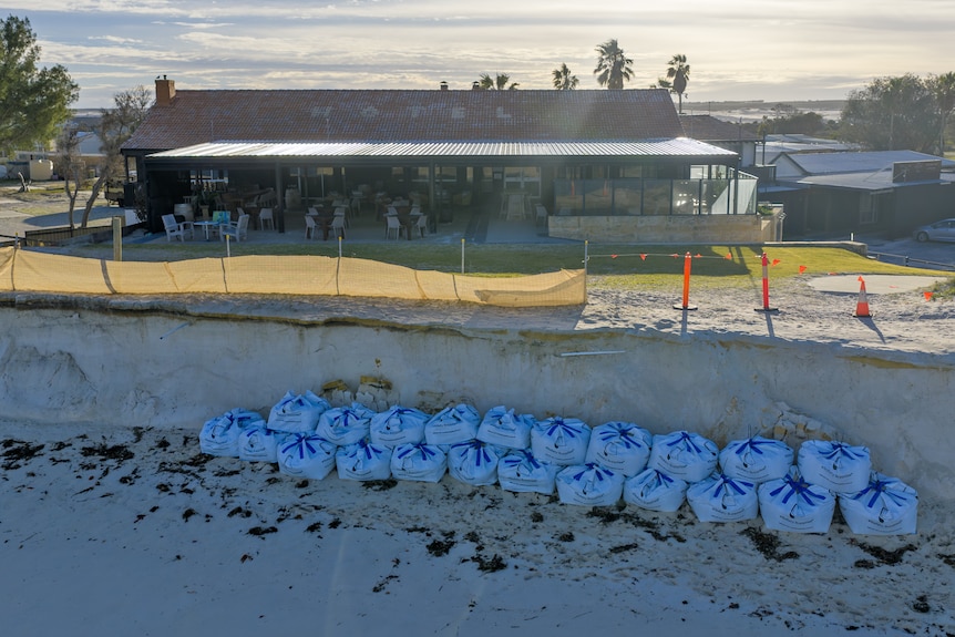 Sand bags sit in front a cliff of sand. A building sits a few metres behind.