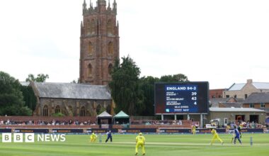 Cricketers play on the grass dressed in yellow and blue kits. A scoreboard shows England 94 for 2. Spectators are sat along the perimiter of the pitch. In the background is a church spire, and other brick buildings against the grey sky.