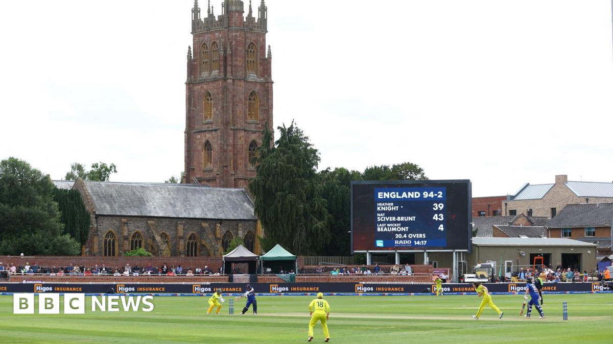 Cricketers play on the grass dressed in yellow and blue kits. A scoreboard shows England 94 for 2. Spectators are sat along the perimiter of the pitch. In the background is a church spire, and other brick buildings against the grey sky.