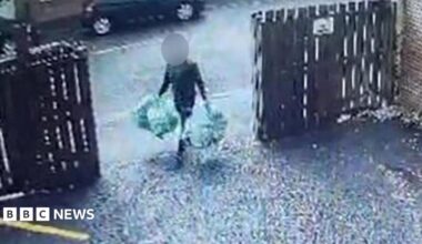 A man carrying four bags of rubbish, two in each hand. He is walking through an open gate into a driveway, with wooden fencing either side. His face is obscured.