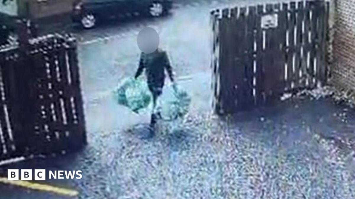 A man carrying four bags of rubbish, two in each hand. He is walking through an open gate into a driveway, with wooden fencing either side. His face is obscured.